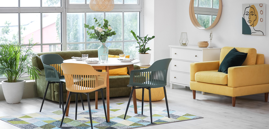 Salle à manger colorée et lumineuse avec canapé en velours vert, fauteuil jaune, table ronde en bois, chaises scandinaves vertes et jaunes avec pieds en métal, tapis à motif graphique, pouf jaune et miroir rond
