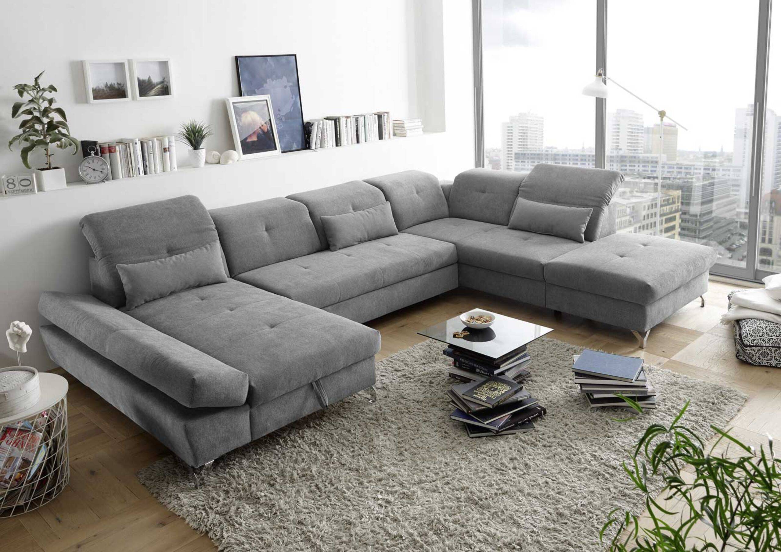 Canapé panoramique gris confortable avec accoudoirs réglables et coffres de rangement, tapis gris, plantes, livres et tableaux 