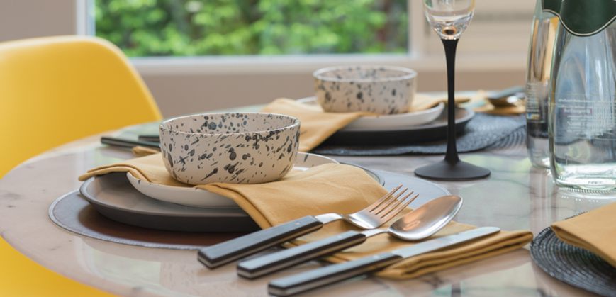 Table à manger ronde en marbre avec assiettes plates grises et blanches, couverts, serviettes jaunse en tissu et bols, flûte à champagne et dessous d’assiette 